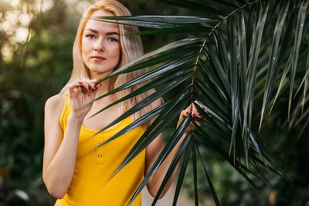 Beautiful girl with natural makeup and blonde hair standing in rainforest among exotic trees. Closeup portrait of magnificent woman in yellow swimsuit among tropical plants. - Фото, зображення