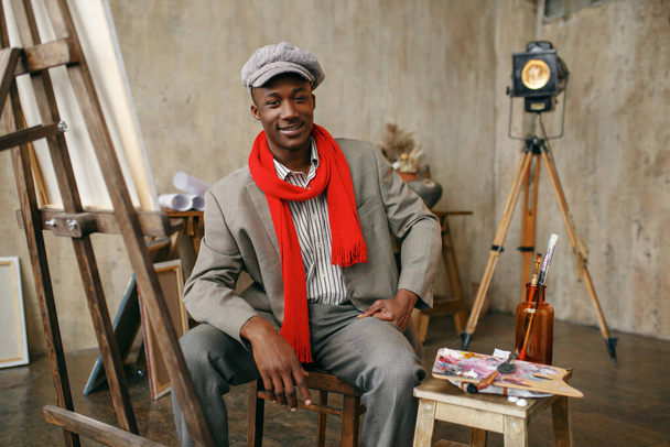 Fashionable male painter in hat and red scarf poses at easel in art studio. Artist draws at his workplace, creative master works in workshop - Фото, зображення