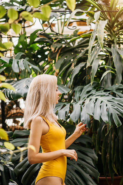 Side view of caucasian young lady stands on background of exotic plants, touches monstera leaf. Profile shot of slim blonde haired woman wearing yellow stylish swimsuit. Nature, vacation. - Valokuva, kuva