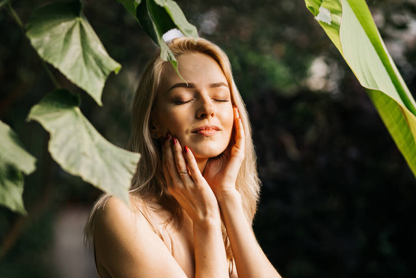 Charming sensual caucasian young woman posing in forest with closed eyes, enjoying summer sun and nature, touching face, using organic cosmetics, applying sunscreen. Skin care, spa concept. - Fotografie, Obrázek