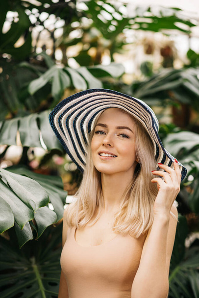 Vertical closeup portrait of happy attractive caucasian young woman with blonde hair, wearing hat, swimsuit, smiling with closed eyes, enjoying sun, relaxing at tropical resort. Summer concept. - Photo, Image