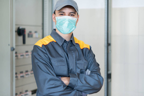 Portrait of an electrician in front of an industrial electric panel in a factory while wearing a mask, coronavirus pandemic concept - Foto, imagen