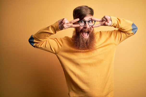 Jóképű ír vörös hajú férfi szakállal szemüveget visel sárga elszigetelt háttér csinál béke szimbólum ujjaival az arcon, mosolygós vidám mutatja győzelem - Fotó, kép