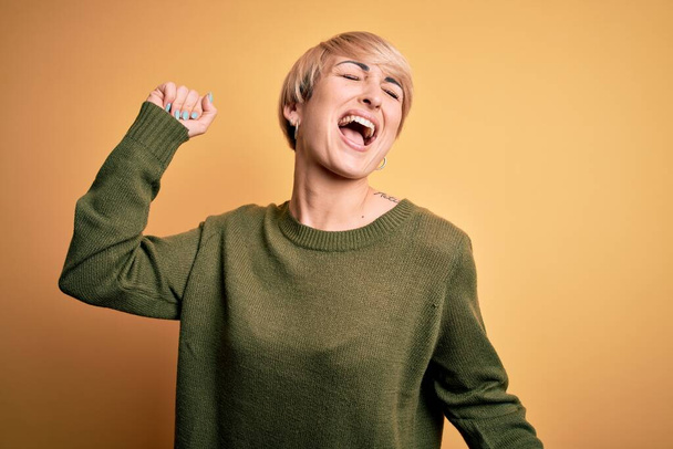 Young blonde woman with modern short hair wearing casual sweater over yellow background Dancing happy and cheerful, smiling moving casual and confident listening to music - Fotó, kép