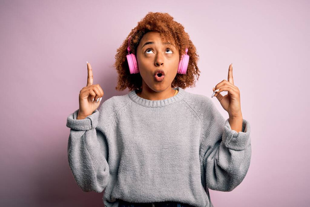 Młoda afro Afroamerykanka z kręconymi włosami słuchająca muzyki za pomocą różowych słuchawek zdumiona i zaskoczona patrząc w górę i wskazując palcami i podniesionymi ramionami. - Zdjęcie, obraz