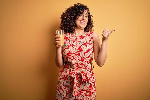 Young beautiful curly arab woman drinking glass of healthy orange juice over yellow background pointing and showing with thumb up to the side with happy face smiling - Photo, image