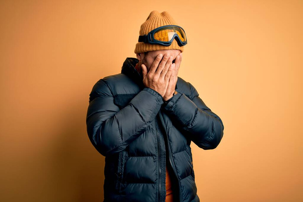 Young handsome skier man with beard wearing snow sportswear and ski goggles with sad expression covering face with hands while crying. Depression concept. - Fotoğraf, Görsel