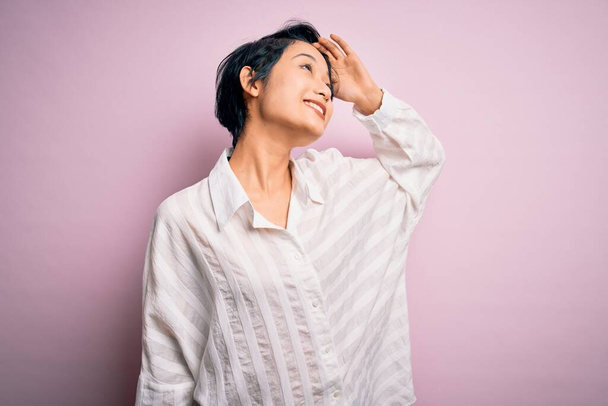 jong mooi aziatisch meisje dragen casual shirt staan over geïsoleerde roze achtergrond glimlachen zelfverzekerd aanraken haar met de hand omhoog gebaar, poseren aantrekkelijk en modieus - Foto, afbeelding
