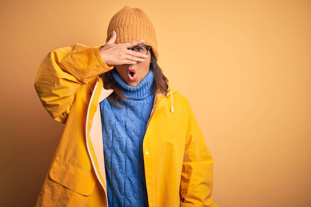 Mujer de mediana edad vistiendo impermeable amarillo y sombrero de invierno sobre fondo aislado mirando con la mano a través de los dedos con expresión avergonzada
. - Foto, imagen