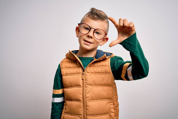 Ein kleines kaukasisches Kind mit blauen Augen, Wintermantel und smarter Brille, lächelnd und selbstbewusst gestikulierend, mit der Hand winzig klein, mit den Fingern schauend und der Kamera. Maßnahmenkonzept. - Foto, Bild