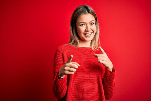 Joven hermosa mujer rubia con suéter casual sobre fondo rojo aislado señalando los dedos a la cámara con la cara feliz y divertida. Buena energía y vibraciones
. - Foto, imagen
