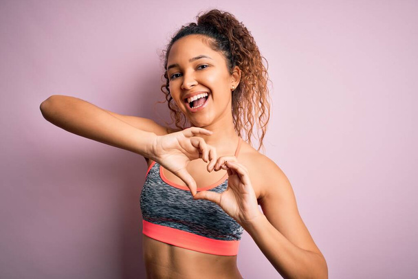 Beautiful sportswoman with curly hair doing sport wearing sportswear over pink background smiling in love doing heart symbol shape with hands. Romantic concept. - Photo, Image