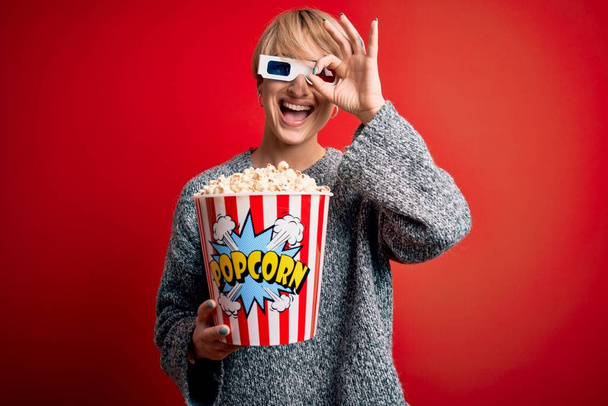 Mujer rubia joven con el pelo corto con gafas de cine 3d y comer palomitas de maíz en el cine con la cara feliz sonriendo haciendo signo bien con la mano en el ojo mirando a través de los dedos
 - Foto, Imagen