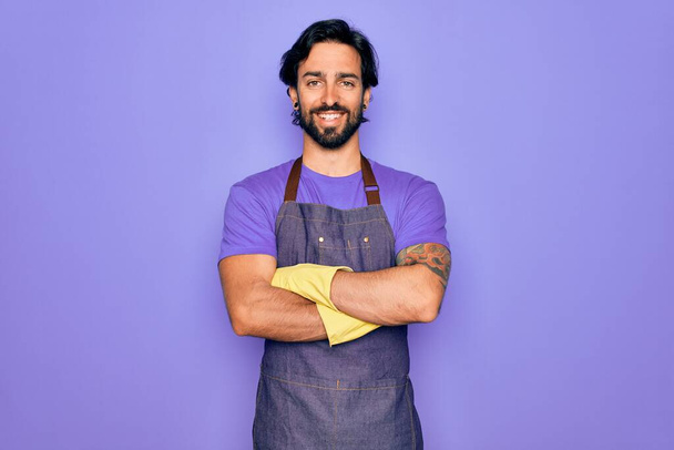 Fiatal jóképű spanyol clenaer férfi házimunka kötényt visel és kesztyűt mos boldog arc mosolyogva keresztbe tett karokkal néz a kamerába. Pozitív személy. - Fotó, kép