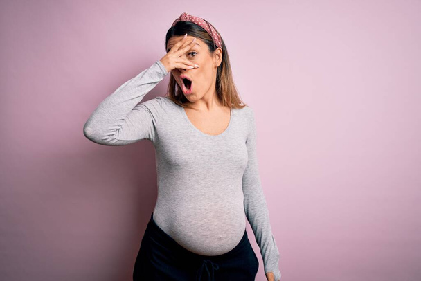 Young beautiful brunette woman pregnant expecting baby over isolated pink background peeking in shock covering face and eyes with hand, looking through fingers with embarrassed expression. - Photo, Image