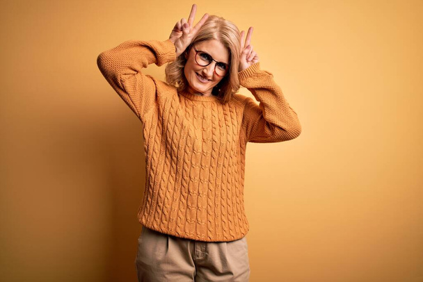 Красивая блондинка средних лет в обычном свитере и очках на жёлтом фоне, изображает смешного и сумасшедшего с пальцами на голове, как кроличьи уши, улыбаясь весело
 - Фото, изображение