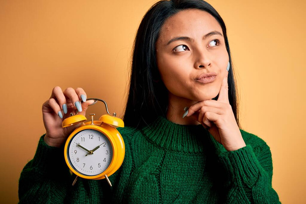 Joven hermosa mujer china sosteniendo reloj despertador vintage sobre fondo amarillo aislado cara seria pensando en la pregunta, idea muy confusa
 - Foto, imagen