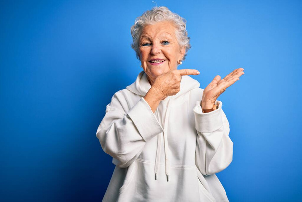 Senior kaunis urheilullinen nainen yllään valkoinen huppari yli eristetty sininen tausta hämmästynyt ja hymyillen kameralle samalla esittää käsi ja osoittaa sormella
. - Valokuva, kuva