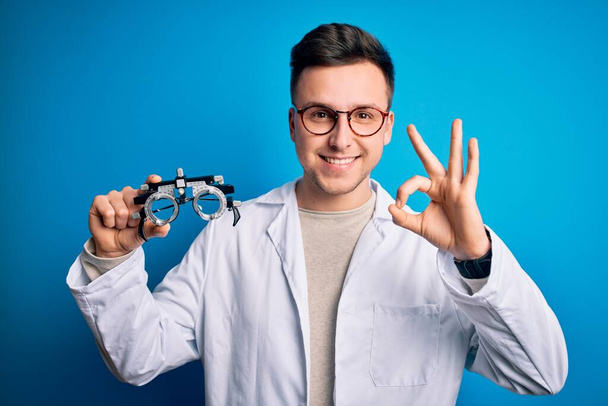 Young handsome professional optic man holding optometrical glasses over blue background doing ok sign with fingers, excellent symbol - Photo, Image