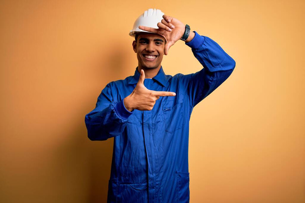 Νεαρός όμορφος Αφροαμερικάνος εργάτης με μπλε στολή και κράνος να χαμογελάει κάνοντας κορνίζα με χέρια και δάχτυλα με χαρούμενο πρόσωπο. Δημιουργία και φωτογραφία έννοια. - Φωτογραφία, εικόνα