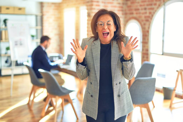 Orta yaşlı, ceketli ve gözlüklü güzel bir iş kadını ofiste çılgın ve çılgın bir başarıyı kollarını kaldırıp heyecanla haykırarak kutluyor. Kazanan konsept - Fotoğraf, Görsel