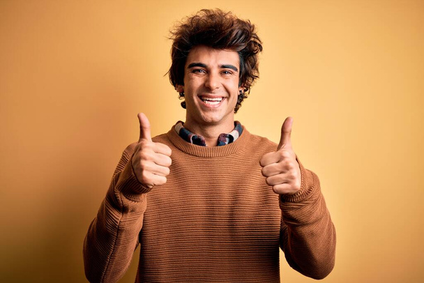 Mladý pohledný muž v neformální košili a svetru přes izolované žluté pozadí znamení úspěchu dělá pozitivní gesto rukou, palce nahoru s úsměvem a šťastný. Veselý výraz a gesto vítěze. - Fotografie, Obrázek