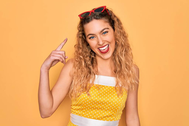 Beautiful blonde woman pin-up with blue eyes wearing red sunglasses over yellow background very happy pointing with hand and finger - Photo, Image