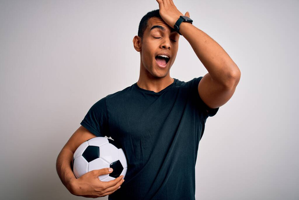 Handsome african american man playing footbal holding soccer ball over white background surprised with hand on head for mistake, remember error. Forgot, bad memory concept. - Foto, imagen