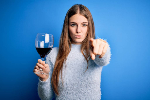 Nuori kaunis punapää nainen juo lasia punaviiniä yli eristetty sininen tausta sormella kameraan ja sinulle, käsi merkki, positiivinen ja luottavainen ele edestä
 - Valokuva, kuva