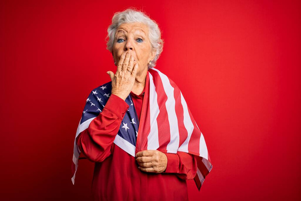 Senior bella dai capelli grigi donna patriottica indossa Stati Uniti bandiera su fondo rosso coprire la bocca con la mano scioccata dalla vergogna per errore, espressione di paura, spaventato in silenzio, concetto segreto
 - Foto, immagini