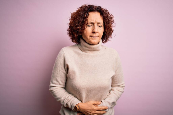 消化不良、痛みを伴う病気が体調不良のため、中年の美しい巻き毛の女性が胃の上に手でピンクの背景にカジュアルなタートルネックセーターを着て。アチェの概念. - 写真・画像