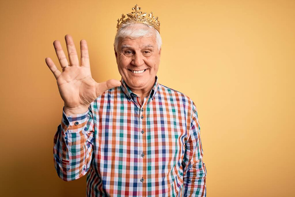 Senior bonito hoary homem vestindo coroa de ouro do rei sobre fundo amarelo isolado mostrando e apontando para cima com os dedos número cinco, enquanto sorrindo confiante e feliz
. - Foto, Imagem