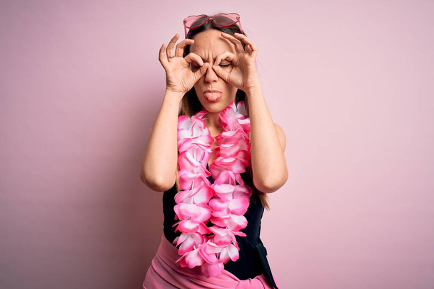Jeune belle femme blonde portant un maillot de bain et un lei hawaïen floral sur fond rose faisant un geste correct comme des jumelles sortant la langue, les yeux regardant à travers les doigts. Expression folle
. - Photo, image