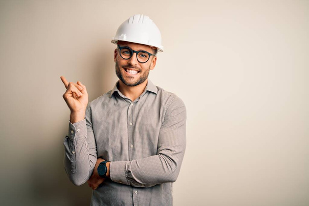 Młody architekt noszący kask ochronny budowniczego na odizolowanym tle z dużym uśmiechem na twarzy, wskazując ręką i palcem w bok patrząc w kamerę. - Zdjęcie, obraz