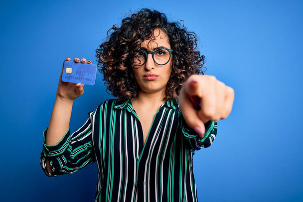 Junge schöne, lockige arabische Frau mit Brille, die Kreditkartengeld zum Bezahlen hält und mit dem Finger in die Kamera und zu Ihnen zeigt, Handzeichen, positive und selbstbewusste Geste von vorne - Foto, Bild