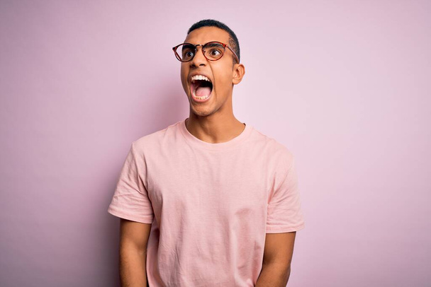 Handsome african american man wearing casual t-shirt and glasses over pink background angry and mad screaming frustrated and furious, shouting with anger. Rage and aggressive concept. - Foto, Imagen