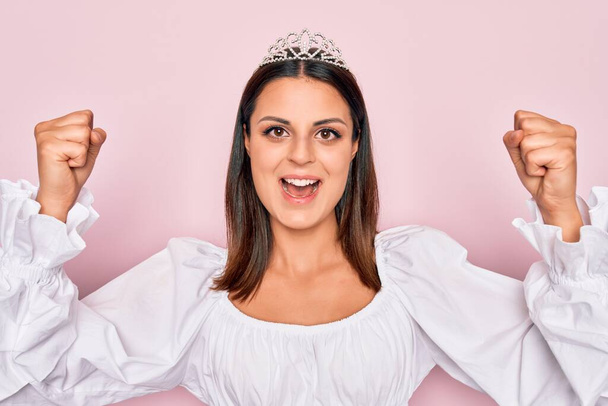 Young beautiful brunette woman wearing princess tiara standing over isolated pink background celebrating surprised and amazed for success with arms raised and open eyes. Winner concept. - 写真・画像