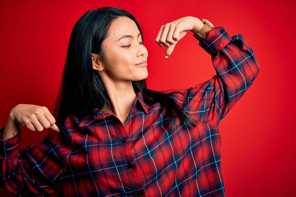 Young beautiful chinese woman wearing casual shirt over isolated red background showing arms muscles smiling proud. Fitness concept. - Fotografie, Obrázek