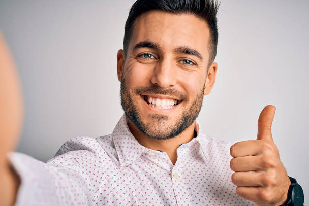 Young handsome man wearing shirt making selfie by the camera over white background happy with big smile doing ok sign, thumb up with fingers, excellent sign - Foto, imagen
