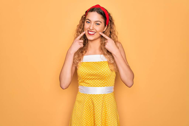 Beautiful blonde pin-up woman with blue eyes wearing diadem standing over yellow background Smiling with open mouth, fingers pointing and forcing cheerful smile - Φωτογραφία, εικόνα