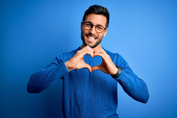 Homem bonito jovem com barba vestindo camisola casual e óculos sobre fundo azul sorrindo no amor fazendo forma de símbolo do coração com as mãos. Conceito romântico
. - Foto, Imagem