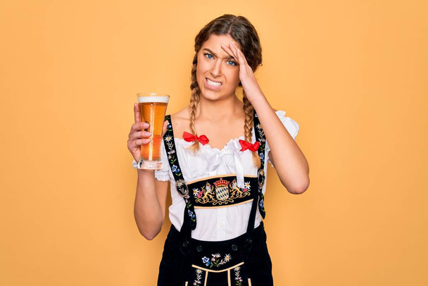 Hermosa mujer rubia alemana con ojos azules vistiendo vestido octoberfest bebiendo vaso de cerveza estresada con la mano en la cabeza, conmocionada con la vergüenza y la cara sorpresa, enojada y frustrada. Miedo y malestar por error
. - Foto, imagen