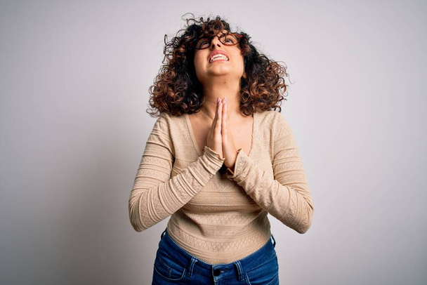 Молодая красивая кудрявая арабская женщина в повседневной футболке и очках на белом фоне просит и молится руками вместе с выражением надежды на лице очень эмоциональным и беспокойным. Умоляю
. - Фото, изображение