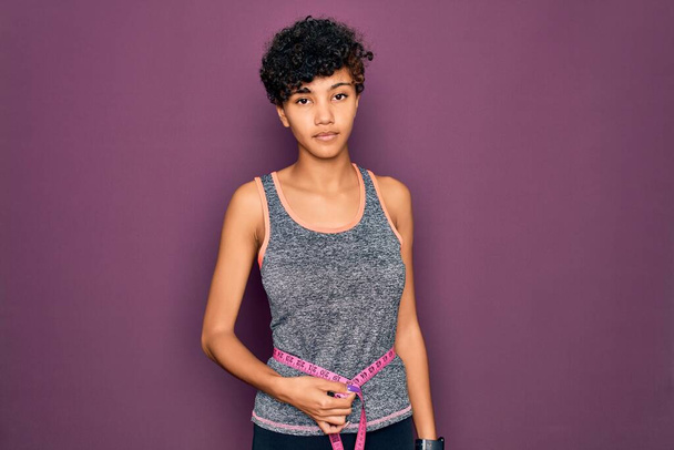 Beautiful african american sporty woman doing exercise controlling weight using tape measure with a confident expression on smart face thinking serious - Foto, Imagen