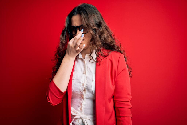 赤い背景の上にジャケットとメガネを着て巻き毛の美しい女性は、鼻の上に指で息を保持し、臭いと嫌な、耐え難い匂いを嗅ぐ。匂いが悪い - 写真・画像
