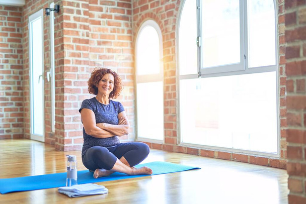 Middle age beautiful sportswoman wearing sportswear sitting on mat practicing yoga at home happy face smiling with crossed arms looking at the camera. Positive person. - Foto, Imagem