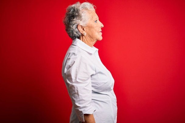 Starsza piękna kobieta w eleganckiej koszuli stojącej nad odizolowanym czerwonym tle patrząc na bok, relaks profil pozować z naturalną twarzą i pewny uśmiech. - Zdjęcie, obraz
