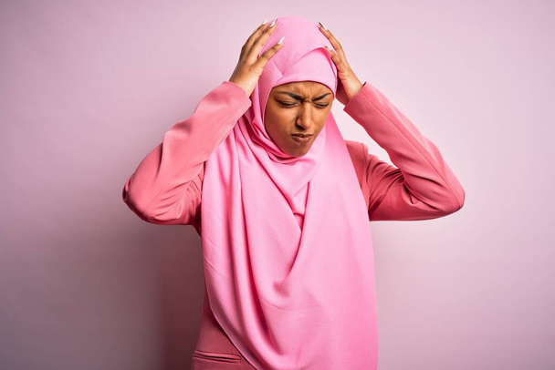Νεαρή Αφρο-Αφρο-Αμερικανίδα που φοράει muslim hijab πάνω από απομονωμένο ροζ φόντο που υποφέρει από πονοκέφαλο απελπισμένη και αγχωμένη επειδή πονάει και ημικρανία. Χέρια στο κεφάλι. - Φωτογραφία, εικόνα