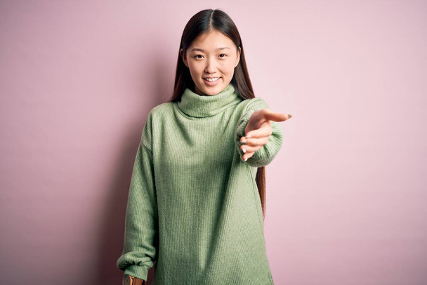 Młoda piękna azjatycka kobieta w zielonym zimowym swetrze na różowym, osłoniętym tle uśmiechnięta, przyjazna, oferująca uścisk dłoni jako powitanie i powitanie. Sprawy zakończone powodzeniem. - Zdjęcie, obraz