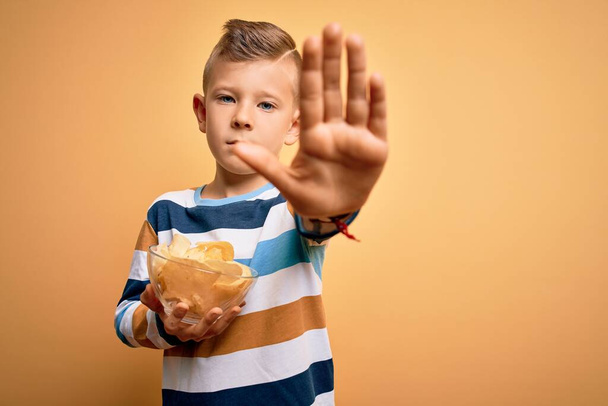 Joven niño caucásico comiendo papas no heatlhy patatas fritas sobre fondo amarillo con la mano abierta haciendo stop sign con expresión seria y confiada, gesto de defensa
 - Foto, Imagen